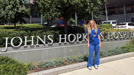 Lene Bleich in blauer medizinischer Kleidung steht vor dem Schild des Johns Hopkins Hospital.